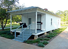Present-day photograph of a whitewashed house, about 15&nbsp;feet wide. Four bannistered steps in the foreground lead up to a roofed porch that holds a swing wide enough for two. The front of the house has a door and a single-paned window. The visible side of the house, about 30&nbsp;feet long, has double-paned windows.